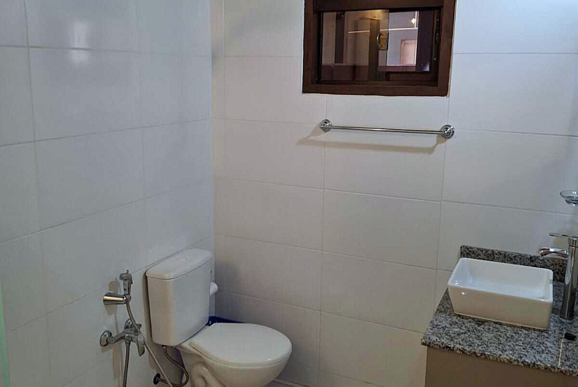 Helles Bad in Paraguay mit weißen Fliesen, Toilette, eckigem Waschbecken auf Granit und kleinem Holzfenster.