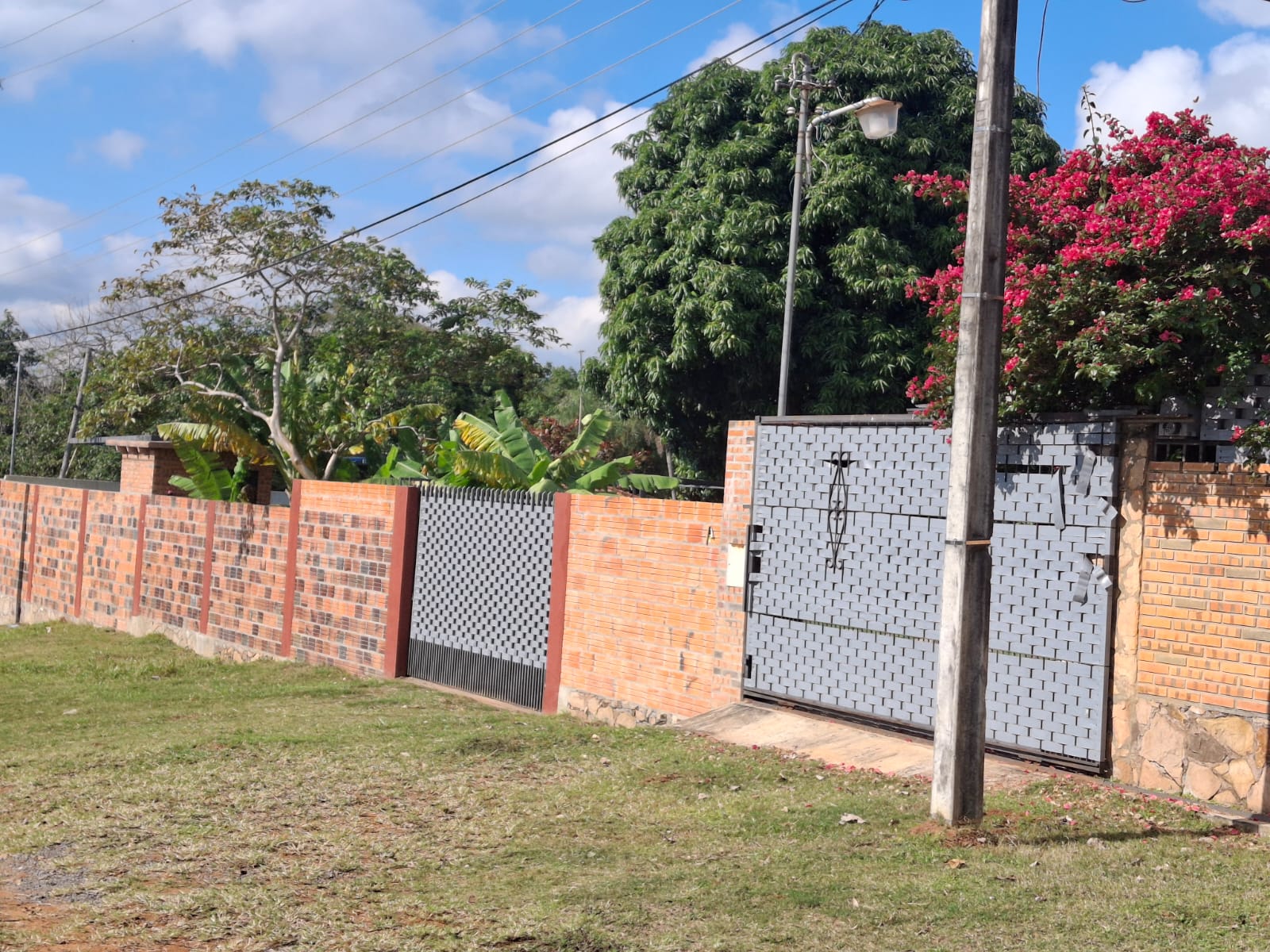 Immobilien Paraguay: Grundstück mit Ziegelmauer, zwei Toren und üppiger tropischer Begrünung unter blauem Himmel.